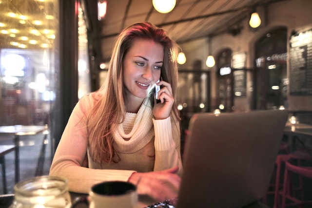 Uma mulher relaxada em um café falando em um smartphone e usando um laptop para procurar serviços de tradução.