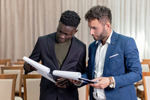 Homens de negócios de terno preto e azul conversando enquanto seguram documentos.