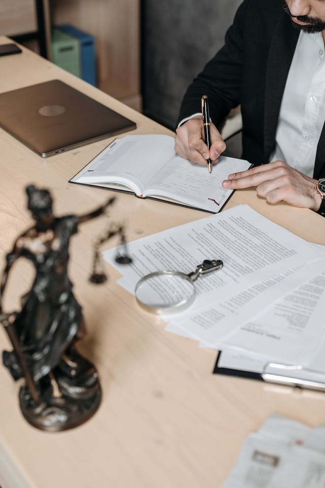 Um homem de terno sentado em uma mesa escrevendo em um caderno enquanto revisa documentos legais para tradução.