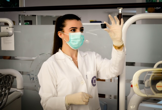 Uma mulher em um laboratório, com jaleco e máscara facial, observa um frasco.