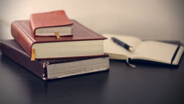 Uma pilha de três livros colocada em uma mesa ao lado de um livro aberto com uma caneta.