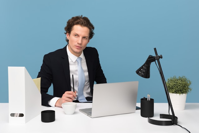 Uma pessoa se senta em uma mesa que tem um laptop, uma xícara de chá, um vaso e outros itens de escritório.