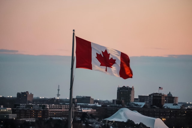 Uma bandeira canadense tremula no céu com um belo pôr do sol ao fundo. 
