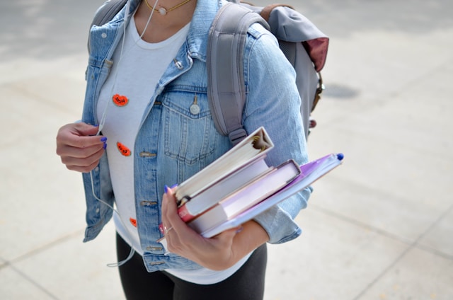 Um estudante no campus segura uma pilha de livros e ouve música com um fone de ouvido. 
