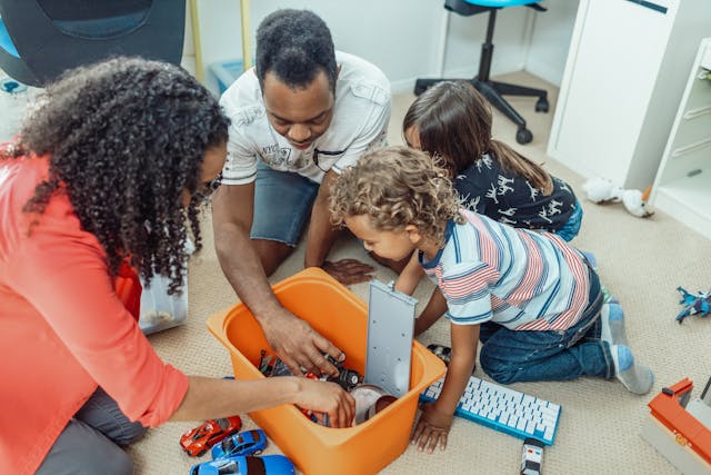 Dois pais adotivos brincam com carrinhos de brinquedo ao lado de seus filhos em seu quarto. 