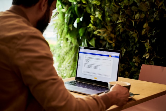 Ein Mann in einer braunen Jacke hält eine Teetasse, während er Texte auf dem Bildschirm eines Laptops betrachtet.
