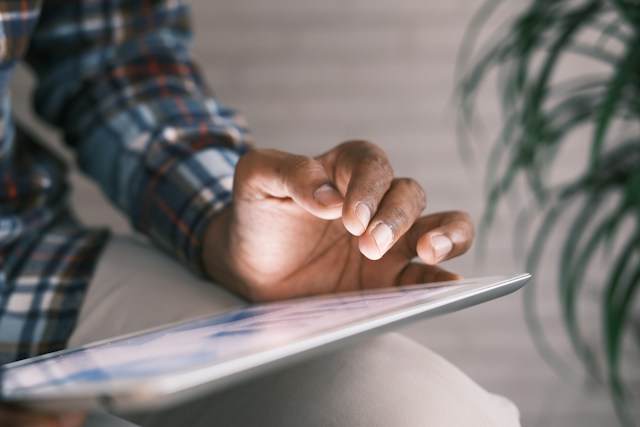 Eine Person in einem blau-weiß karierten langärmeligen Hemd hält ein silbernes digitales Tablet.
