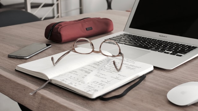 Ein aufgeklappter Laptop und ein Notizblock mit Gläsern liegen auf einem Holztisch.