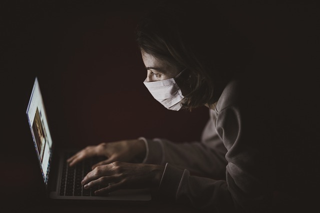 Eine Person, die eine Nasenmaske trägt, benutzt einen Laptop in einer dunklen Umgebung.
