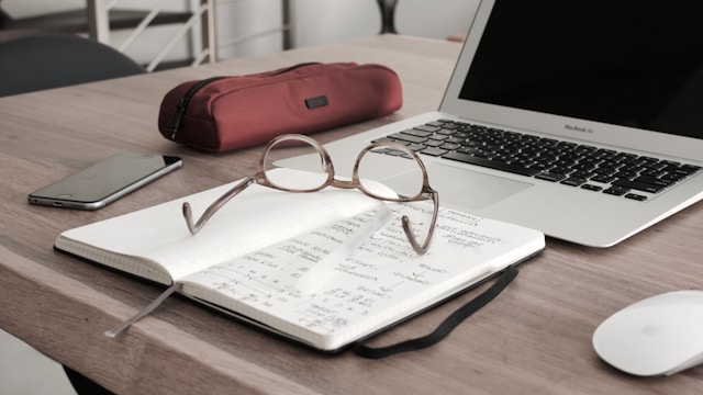 Un ordenador portátil abierto y un bloc de notas con gafas colocados sobre una mesa de madera.