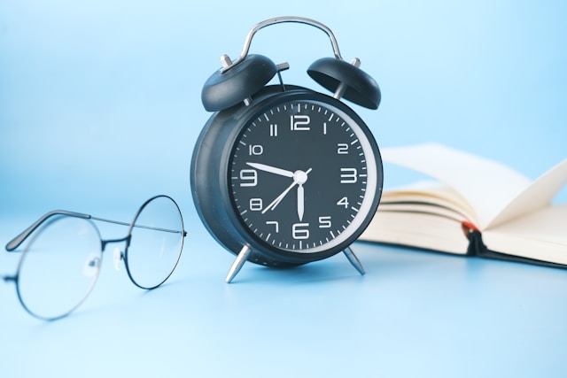 Un despertador negro con unas gafas y un libro al lado.
