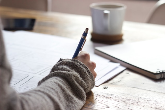 Una persona escribe en un papel blanco sobre una mesa de madera con una taza de café sobre la mesa.
