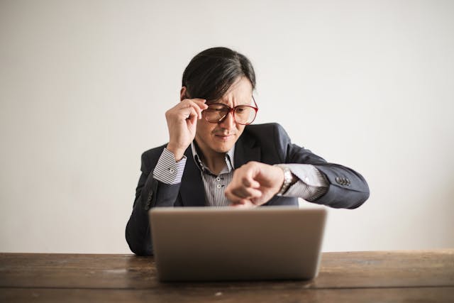 Un homme en costume et lunettes regarde l'heure sur sa montre-bracelet.
