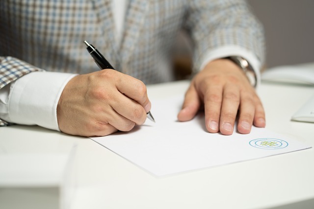 Une personne vêtue d'un costume à carreaux bleus et bruns signe un document à l'aide d'un stylo.