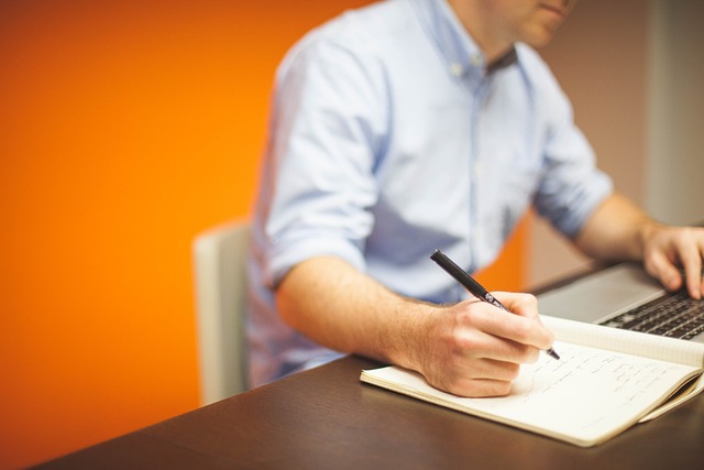 Une personne en chemise bleue écrit dans un livre tout en utilisant un ordinateur portable.