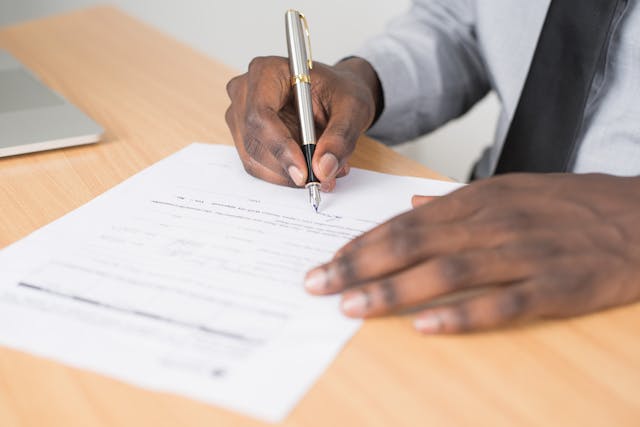 Une personne en chemise blanche et cravate signe un document sur une table.
