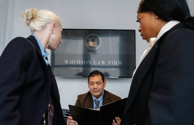 Un avocat lit un document sous le regard de deux autres personnes.
