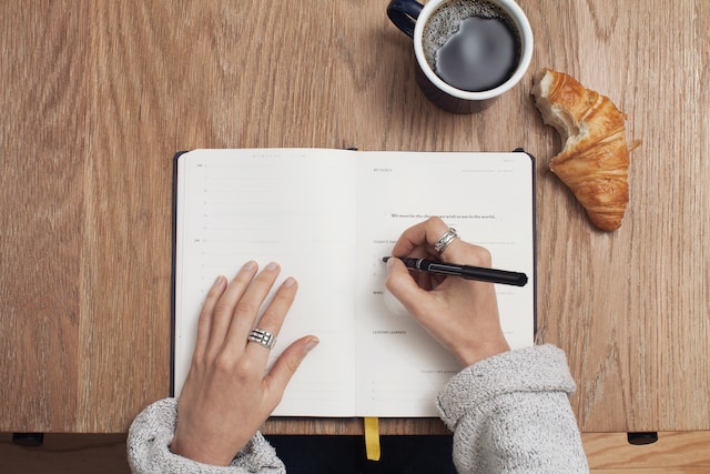 Un'immagine di qualcuno che scrive in un libro su un tavolo con accanto un biscotto e una tazza.