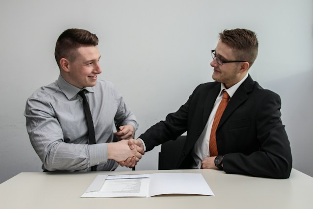 Due uomini si fronteggiano mentre si stringono la mano in segno di accordo su un tavolo che contiene un documento.
