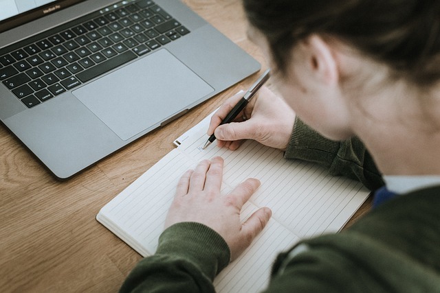 Una persona scrive in un libro davanti a un laptop d'argento.
