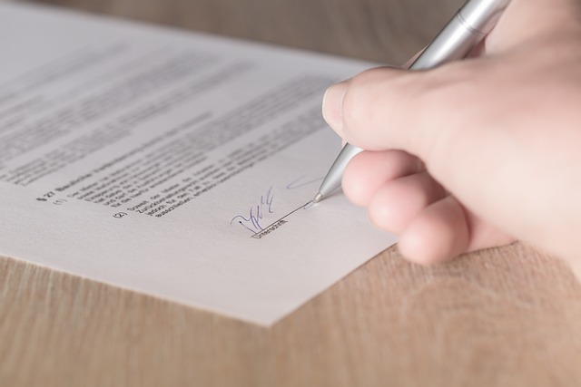 Una mano usa una penna d'argento per firmare su carta stampata su una superficie di legno.

