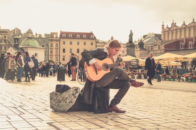 Osoba siedzi i gra na gitarze na środku ulicy wypełnionej ludźmi.
