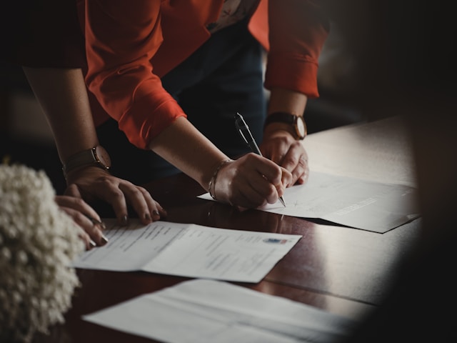 A person in an orange long-sleeve shirt writing on paper on a wooden table.
