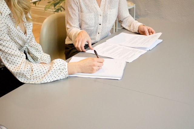 Two faceless women working on documents and writing in a notebook while seated at a desk.