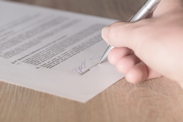 A hand uses a silver pen to sign on printed paper on a wooden surface.
