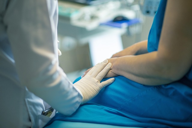 A person in a white lab coat places their gloved hand on the lap of a person wearing a blue hospital gown.
