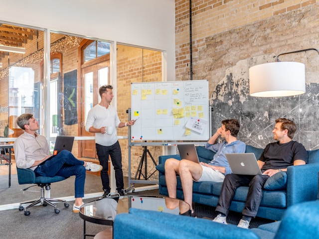 A group of people watches as a man presents ideas and strategies on a whiteboard.
