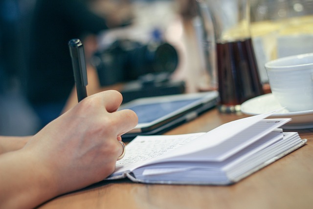 A hand writes in a book on a wooden surface beside a white ceramic teacup.
