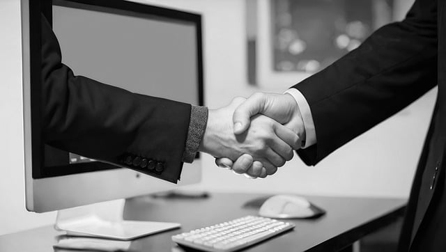 Two people wearing suits shake hands before a computer.
