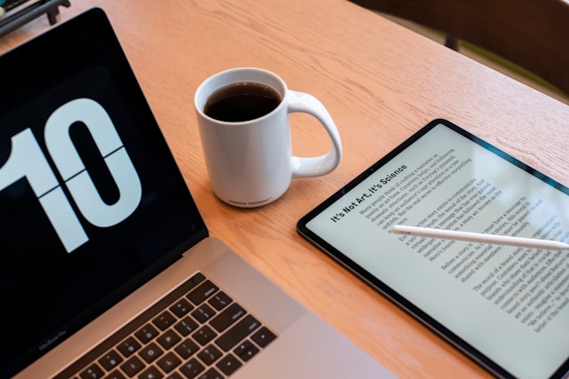 A tablet on a table beside a laptop and coffee cup displays a digital Word file.