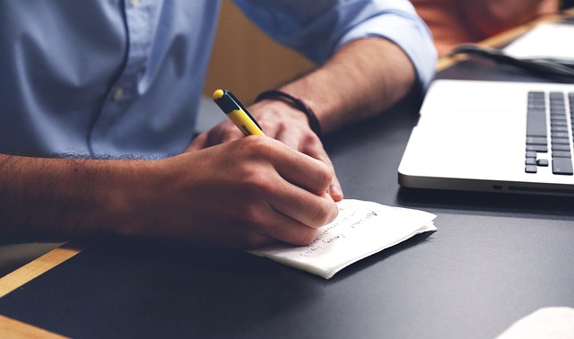 A person with a blue shirt writes on paper before a silver laptop.