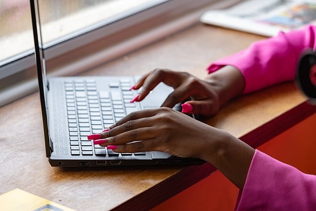 A person with a pink shirt types on a laptop on a wooden surface.