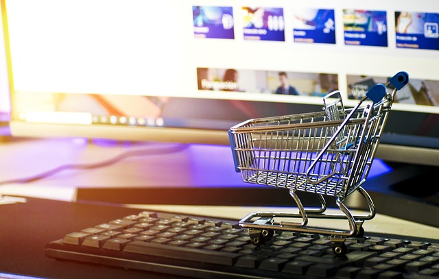 A miniature shopping cart is on a black computer keyboard with a shopping website in the background.
