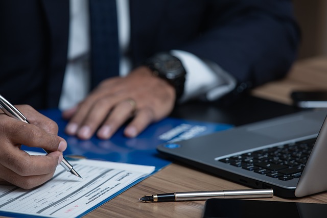 A person signs a document on a desk in front of a laptop.
