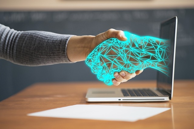 A human is in a handshake with a robotic hand coming out of a laptop.
