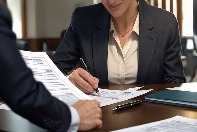 A person in a black suit writes in a document while sitting across another person.
