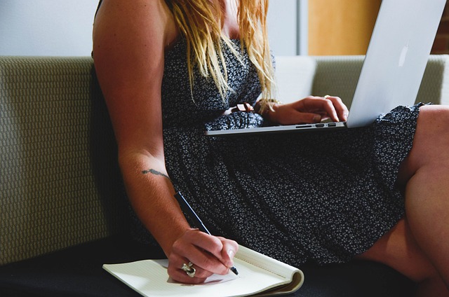 A person places a silver laptop on their leg and writes in a book while sitting on a sofa.
