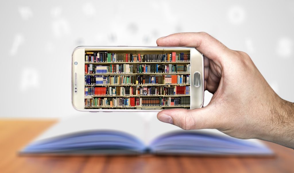 A hand holds a white smartphone displaying a bookshelf with an open book in the background.
