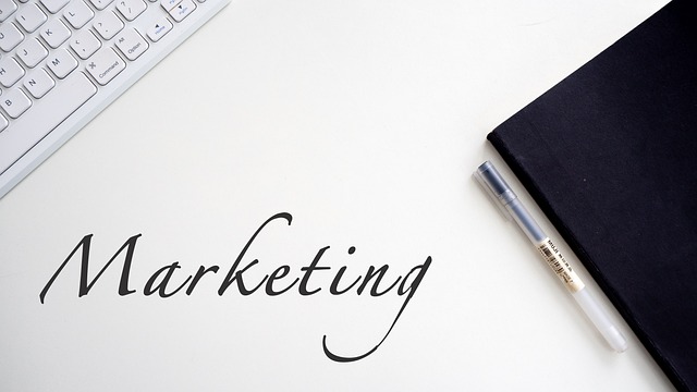 The word “Marketing” between a pen, a white keyboard, and a black notepad on a white surface.
