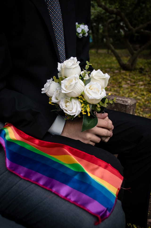 A close-up of a groom holding a white rose wedding bouquet next to a rainbow flag. 
